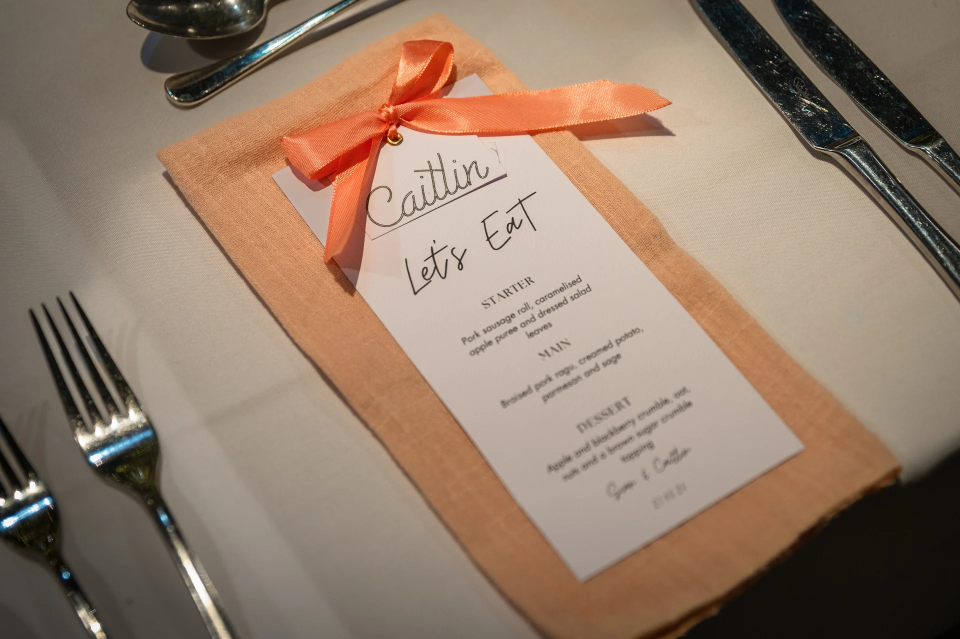 An elegant wedding place setting features a peach napkin tied with a matching ribbon and a personalised menu card resting on a white tablecloth.