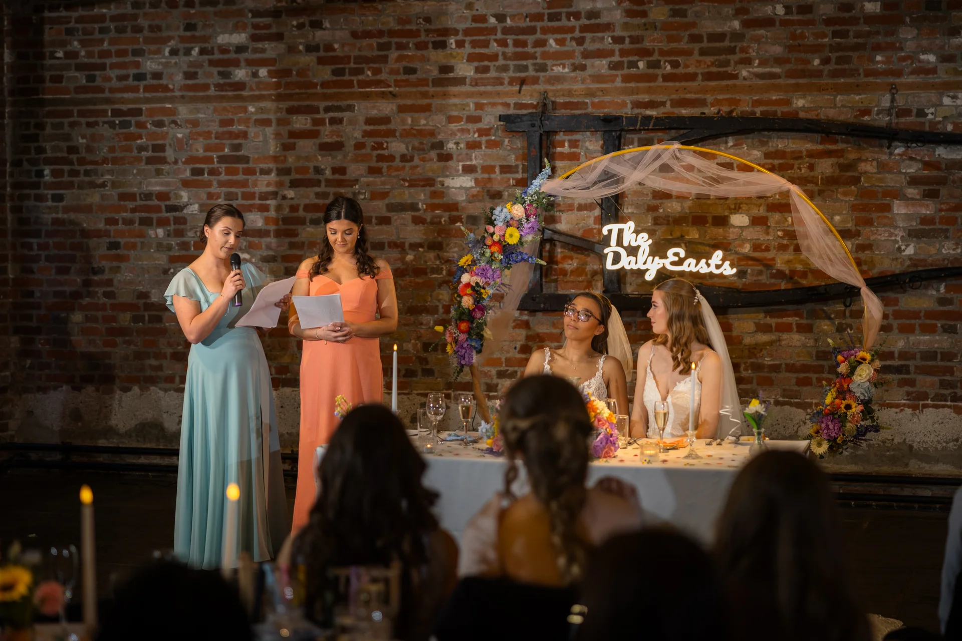 Two bridesmaids deliver speeches standing next to the top table where two brides are seated in front of an exposed brick wall and a decorative neon sign.