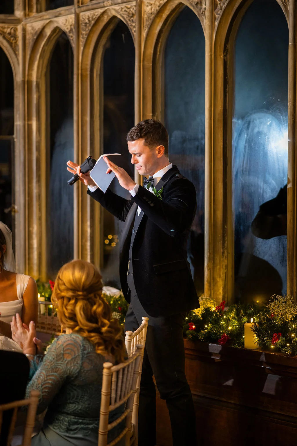 A groom delivers a speech in a historic castle venue featuring dramatic stone arch windows and candlelit tablescapes.