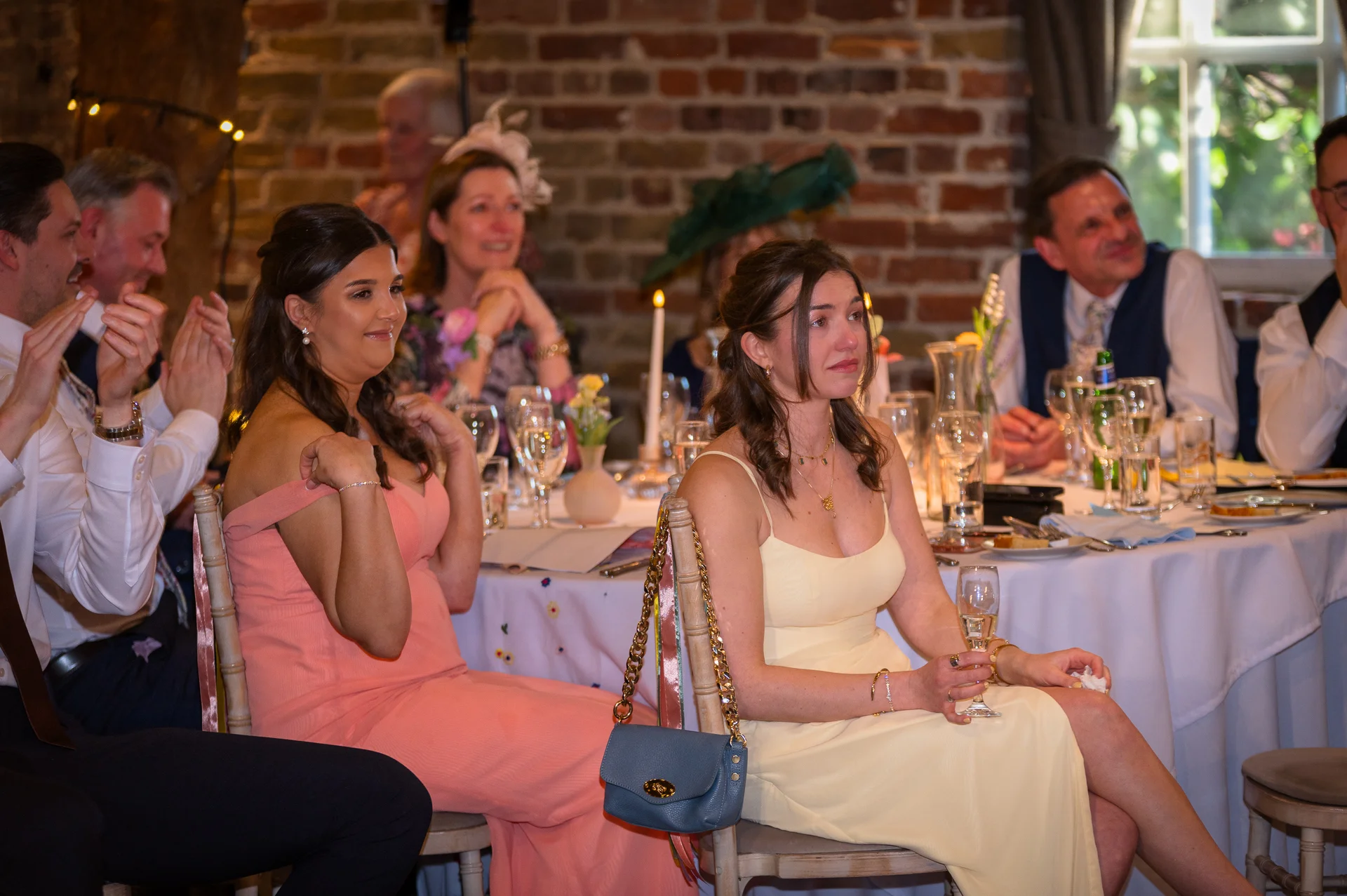 Emotional wedding guests are seated at a round table during speeches, with one guest holding back tears while others listen attentively.