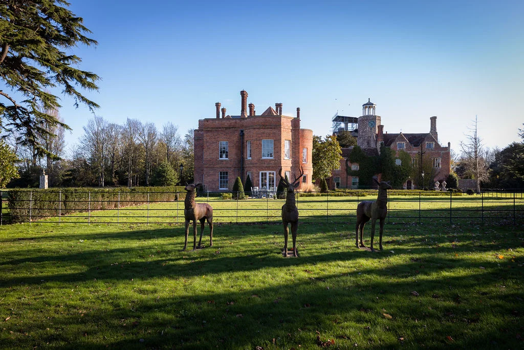 A wide exterior shot of a grand red brick country house under a clear blue sky, featuring manicured lawns and decorative deer statues in the foreground.