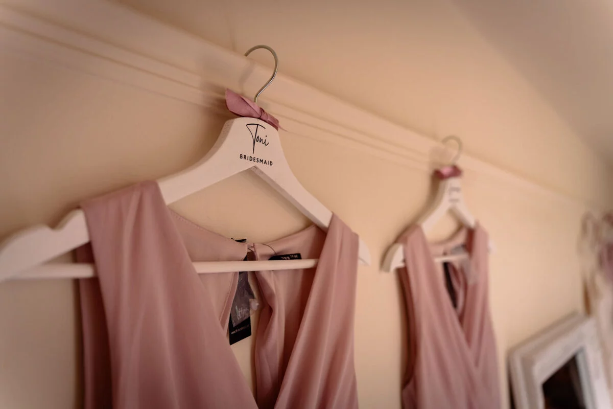 Two pink bridesmaid dresses hang neatly on personalised white wooden hangers against a neutral wall during morning preparations.