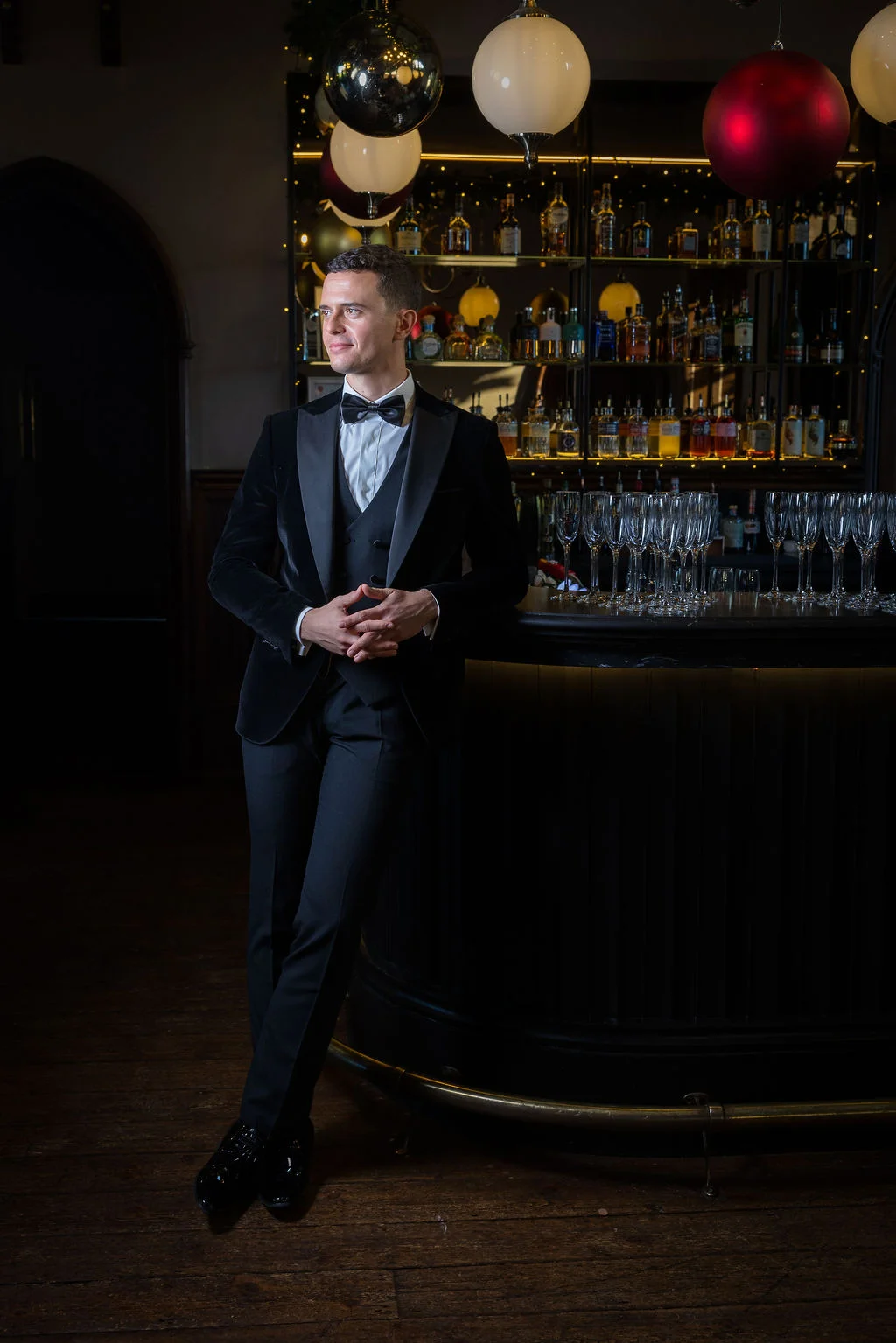 A handsome groom in a black velvet tuxedo leans against a sophisticated pub bar stocked with bottles and crystal glassware.
