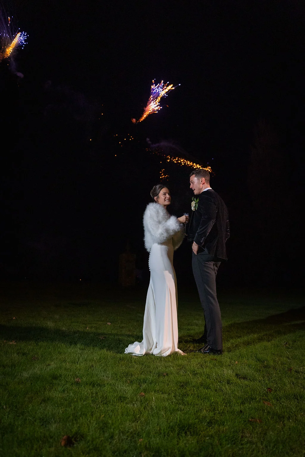 A newlywed couple stands outdoors at night on a lawn, laughing together under a bright display of fireworks against the dark sky.