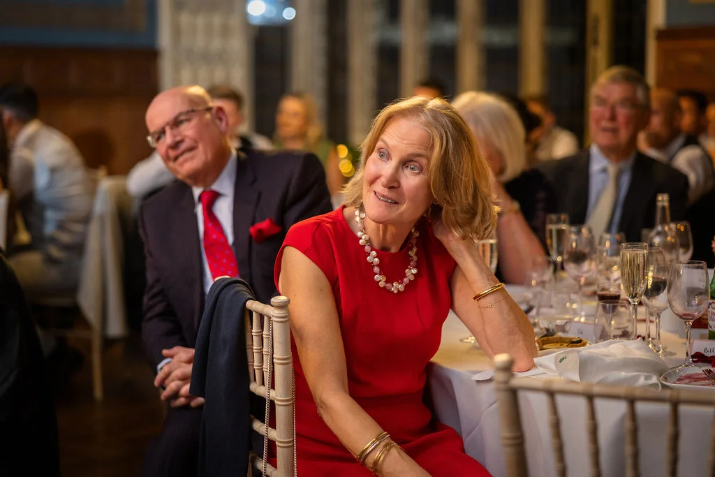 A mature female wedding guest in a red dress sits attentively at a banquet table during speeches, smiling as she listens.