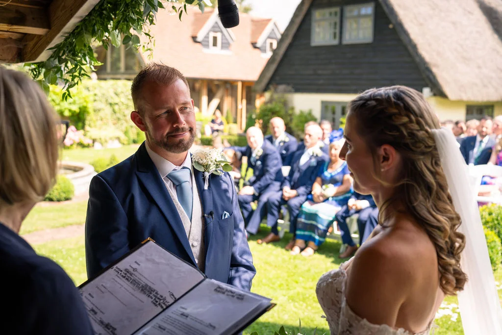 An outdoor wedding ceremony takes place in a sunny garden as the groom smiles lovingly at the bride while standing beside the celebrant holding a register.