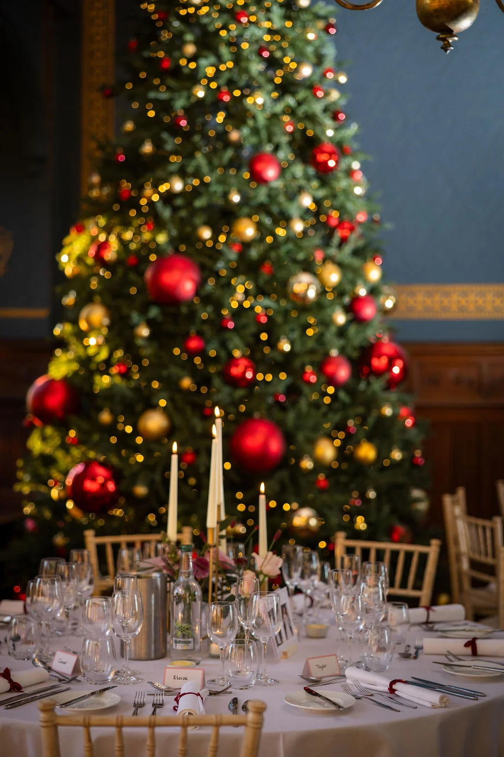 A beautifully styled wedding reception table features delicate place settings, glowing taper candles, and fine glassware set against a large, beautifully decorated Christmas tree backdrop.