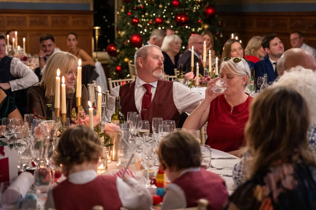 Wedding guests enjoy a festive wedding breakfast in a candlelit, wood-panelled room with a large Christmas tree in the background.