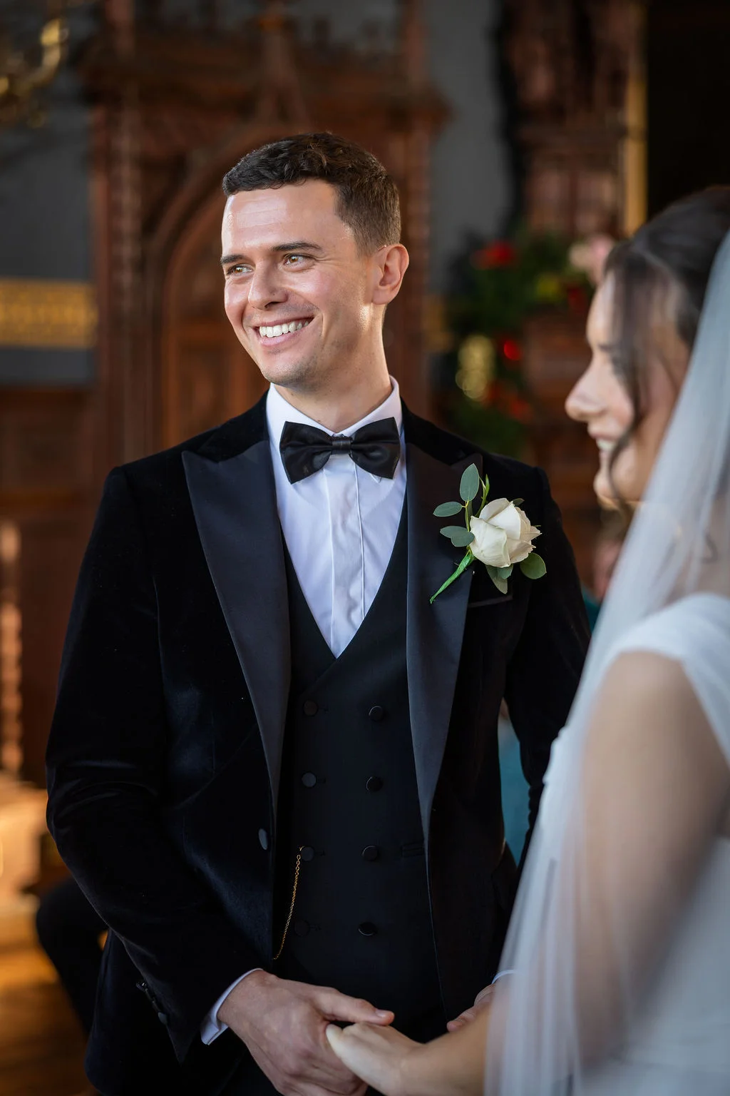 A smiling groom in a black velvet tuxedo with a white rose buttonhole holds hands with his bride during their wedding ceremony.