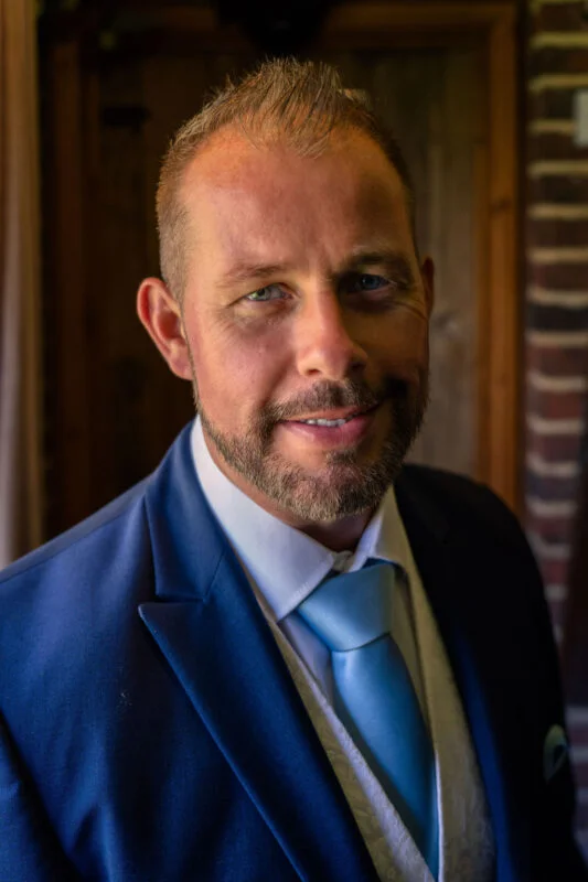 A close-up portrait of a smiling groom dressed in a dark suit with a light blue tie and waistcoat.