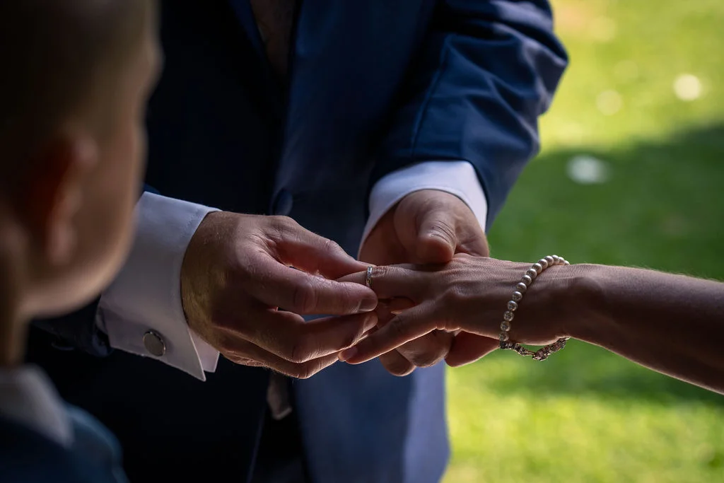 A close-up shot captures the moment a groom in a navy suit places a wedding ring onto the bride's finger, adorned with a pearl bracelet.