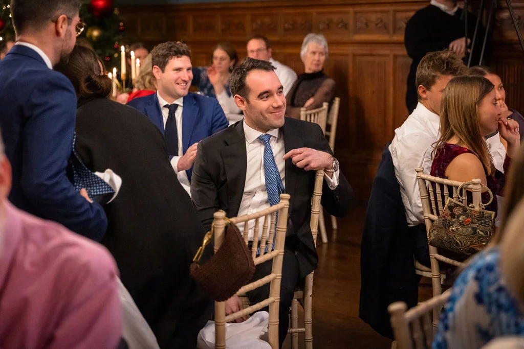 A guest smiles and points towards someone during the wedding breakfast in a wood-panelled room with seated guests around tables.