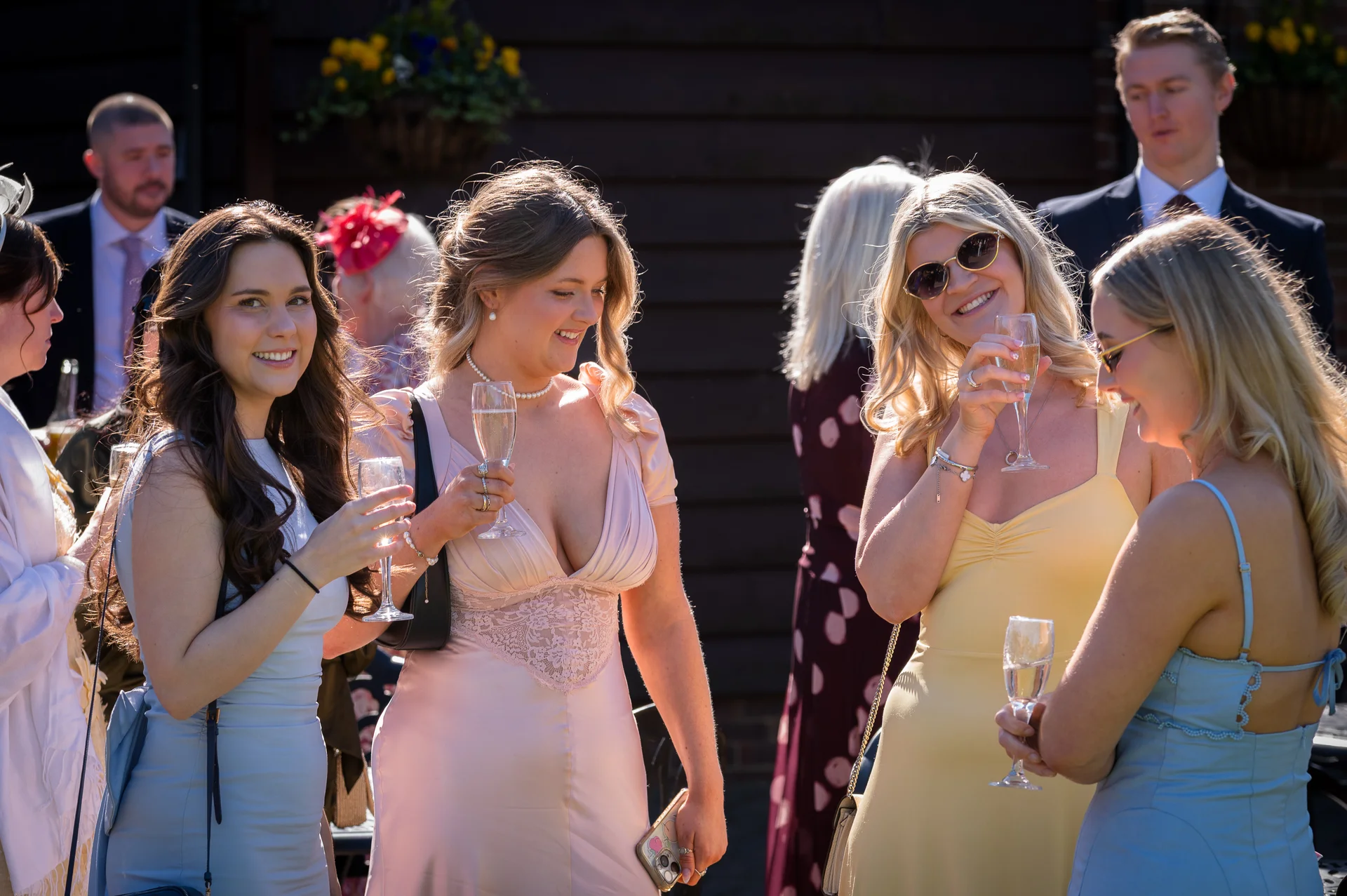 A group of wedding guests mingle and share drinks outdoors in the bright sunshine, holding champagne flutes and laughing together.