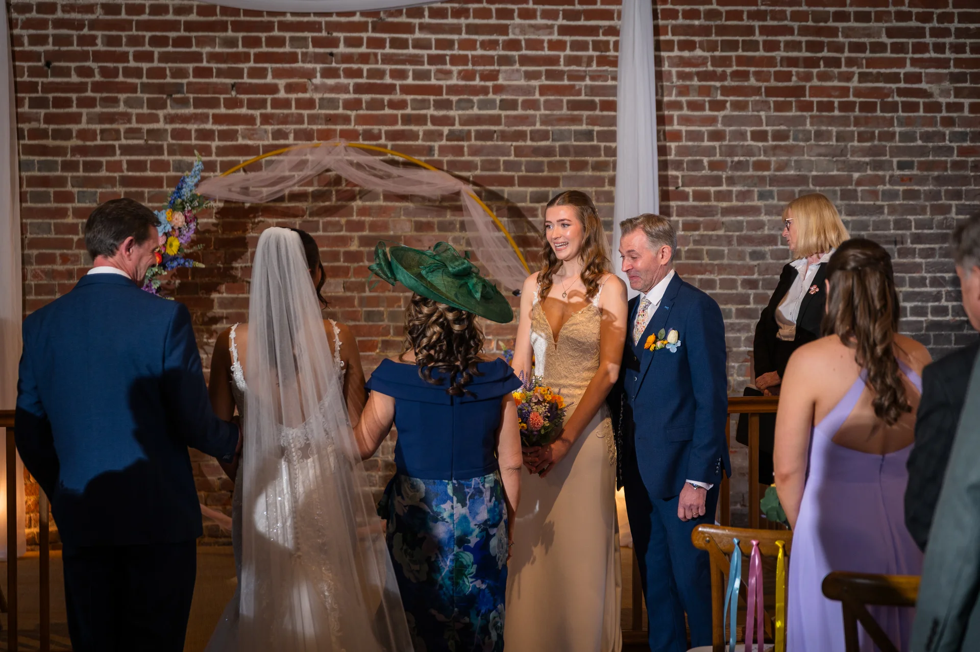 A bride walks down the aisle accompanied by her father, facing the groom and wedding party in an indoor venue with exposed brick walls and a floral arch.