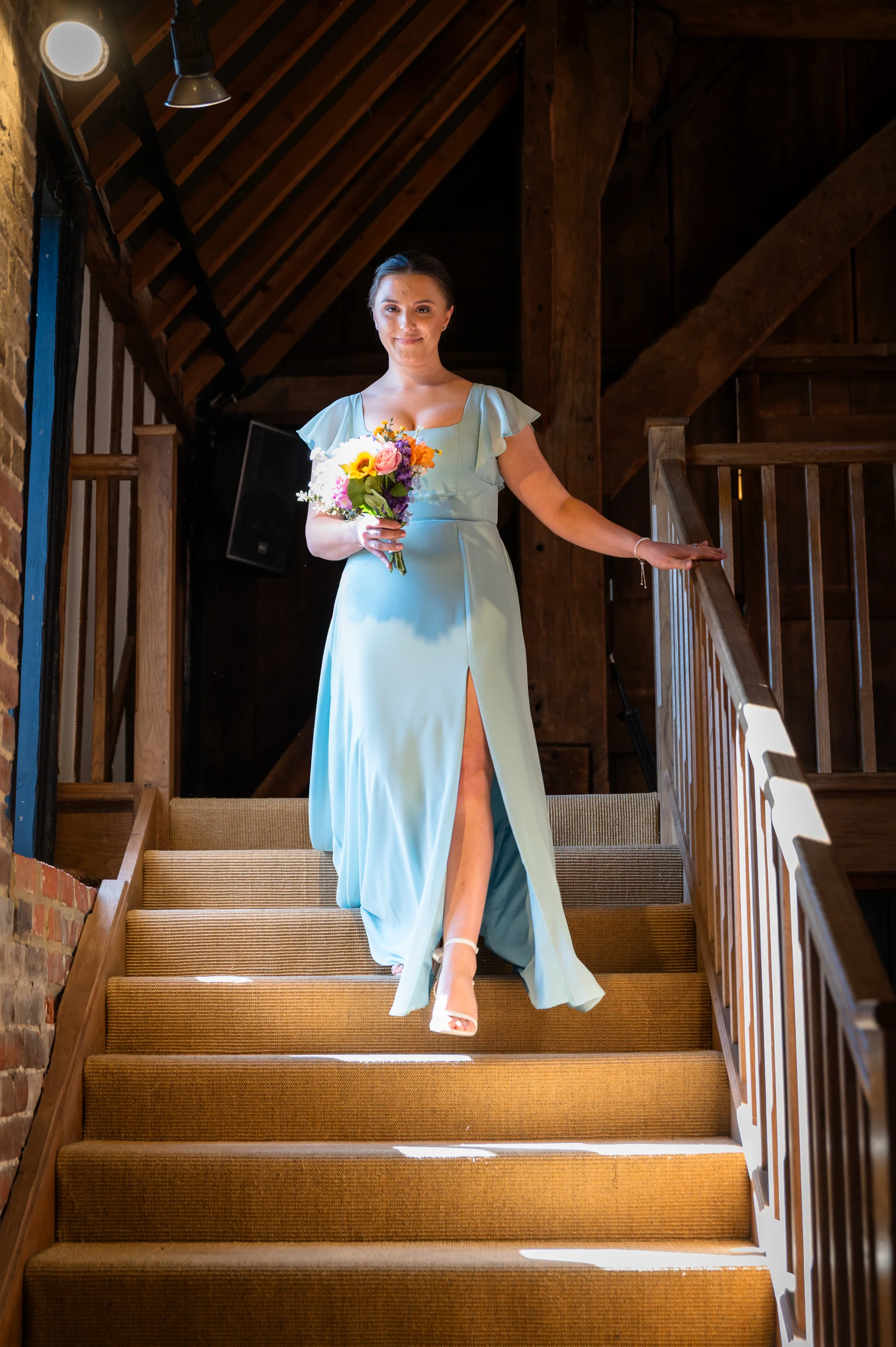 A bridesmaid in a pale blue dress with flutter sleeves and a thigh-high split stands on a wooden barn staircase, holding a colourful bouquet.