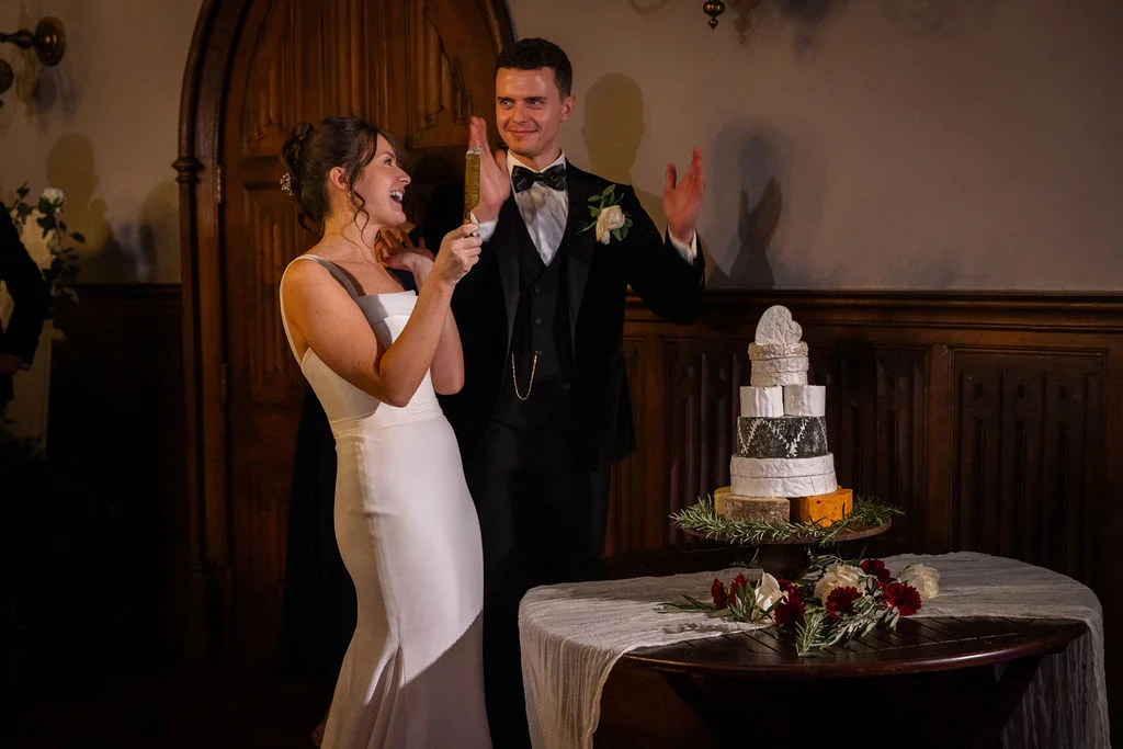 A joyful bride and groom laugh and clap beside a tiered cheese tower wedding cake in a wood-panelled historic venue.