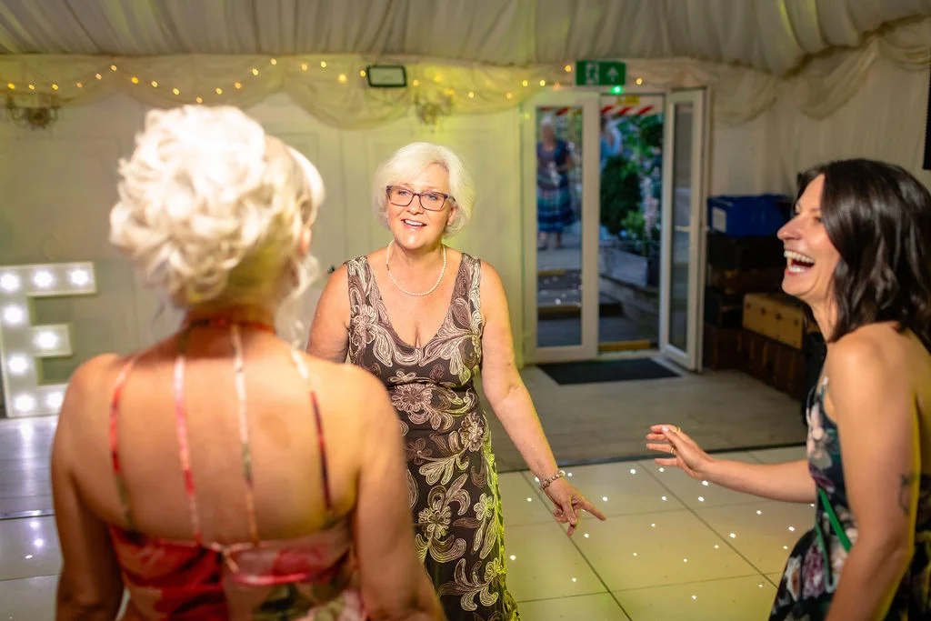 Three female guests laugh and dance joyfully together on an illuminated dance floor inside a fabric-draped marquee.