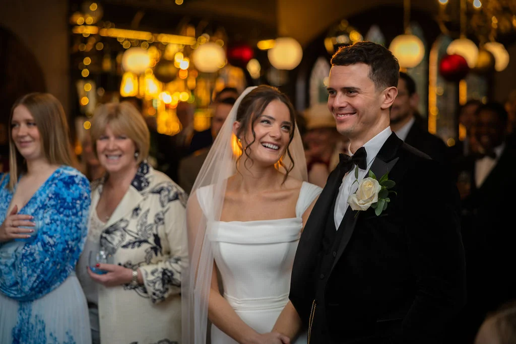 A joyful bride and groom share a tender, laughing glance during their wedding reception while surrounded by smiling guests.