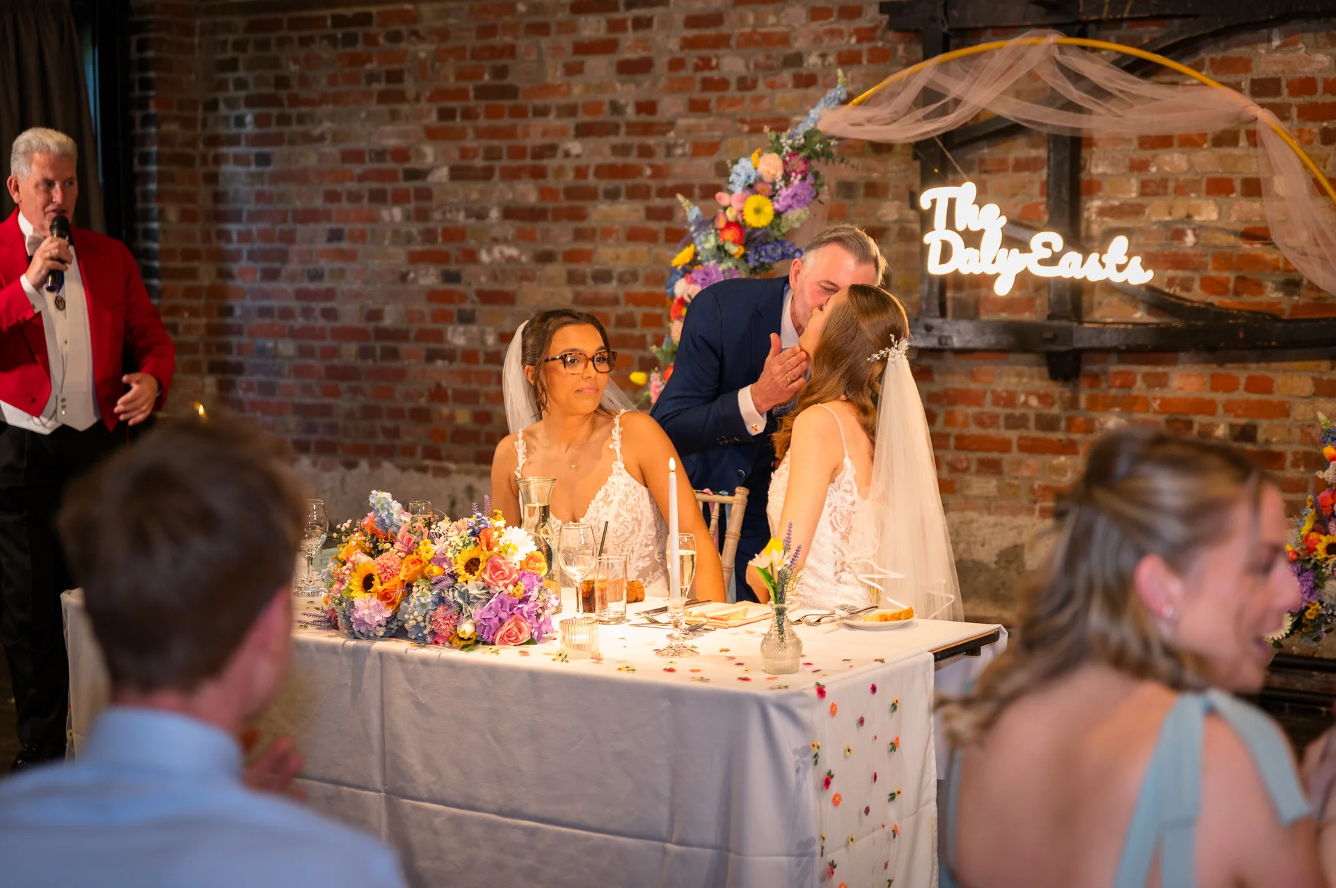 A same-sex couple shares a tender kiss at the top table during wedding speeches, set against an exposed brick wall with colorful floral installations and a custom neon sign.