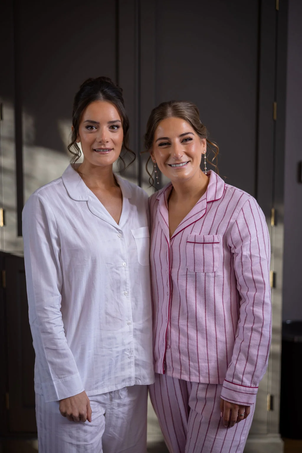 Two smiling women standing indoors while wearing matching striped pyjamas during bridal preparations.