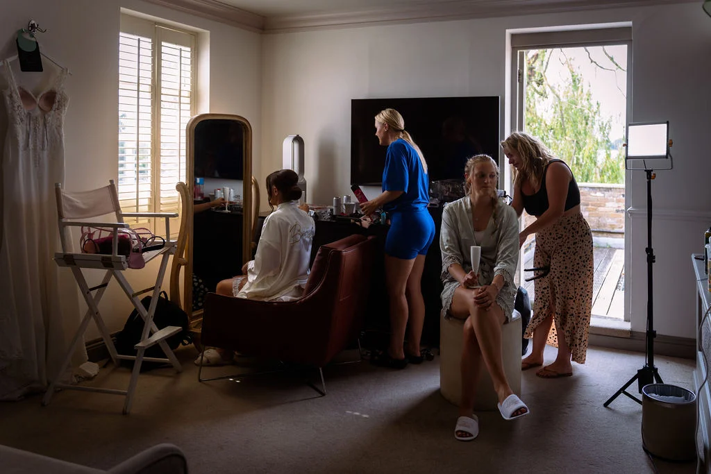 The bride sits before a mirror in a white robe while bridesmaids relax and prepare in a bright hotel room during morning bridal preparations.