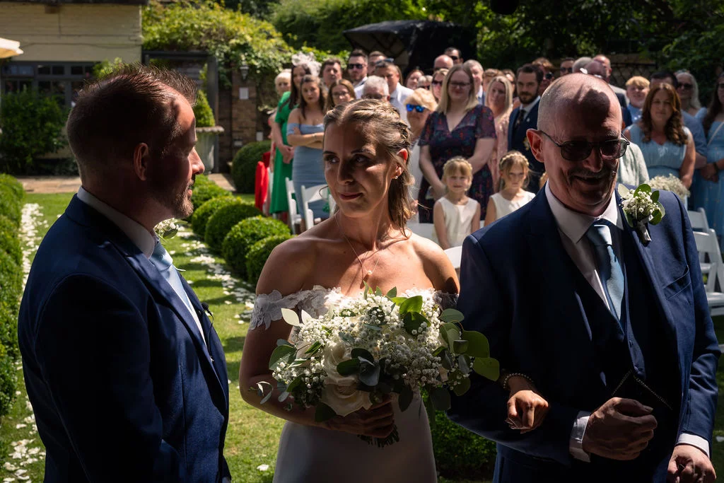 The bride walks down an outdoor garden aisle accompanied by her father, holding a white rose and baby's breath bouquet towards the waiting groom.