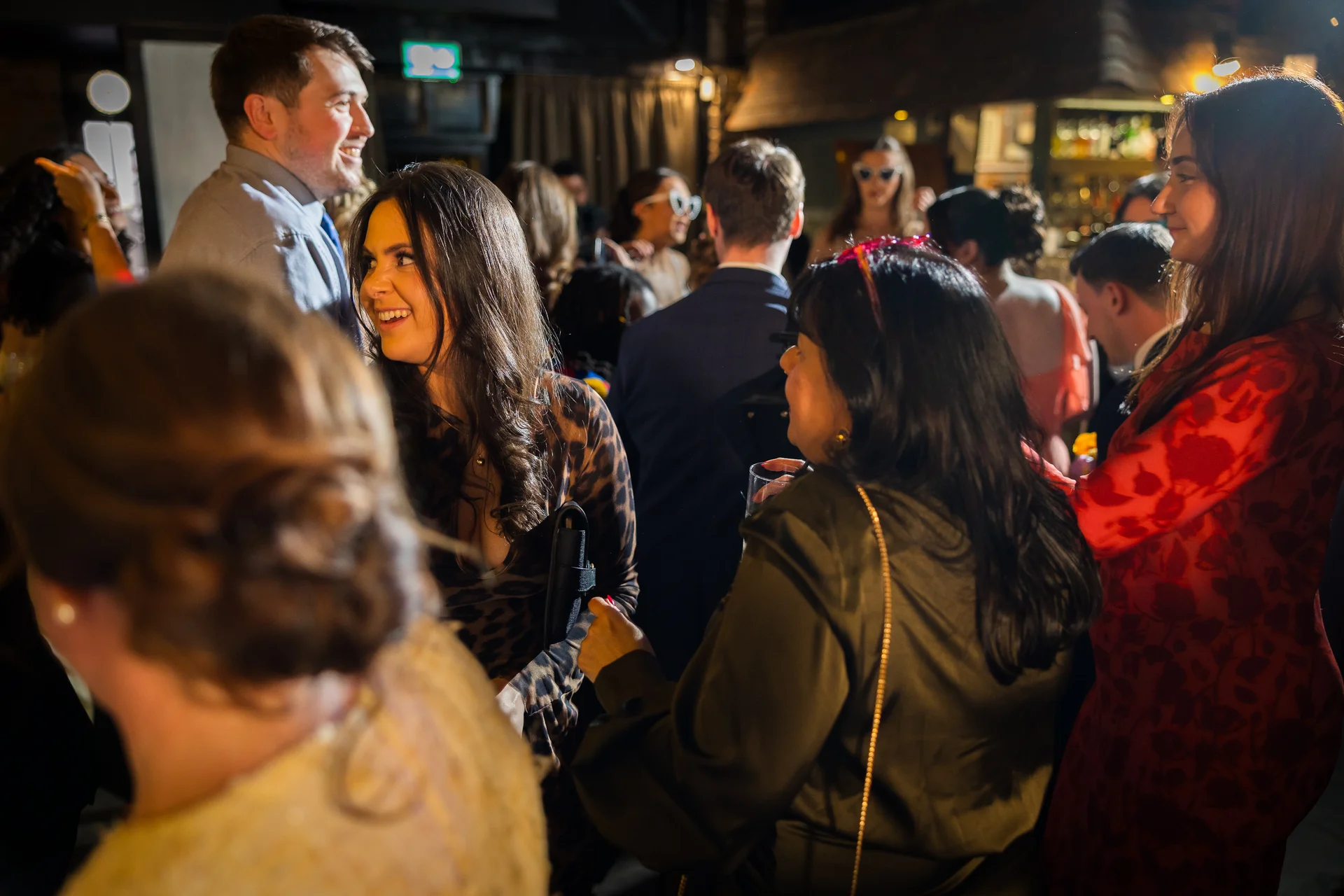 Candid shot of wedding guests laughing and mingling together inside a lively evening reception venue under warm indoor lighting.