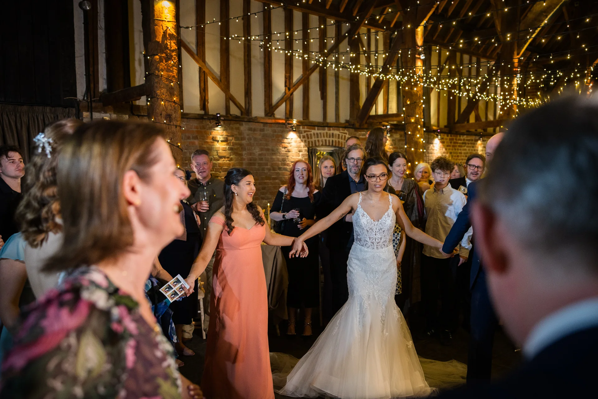 A bride in a strapless lace fit-and-flare gown dances in a circle with guests inside a rustic barn illuminated by a canopy of fairy lights.