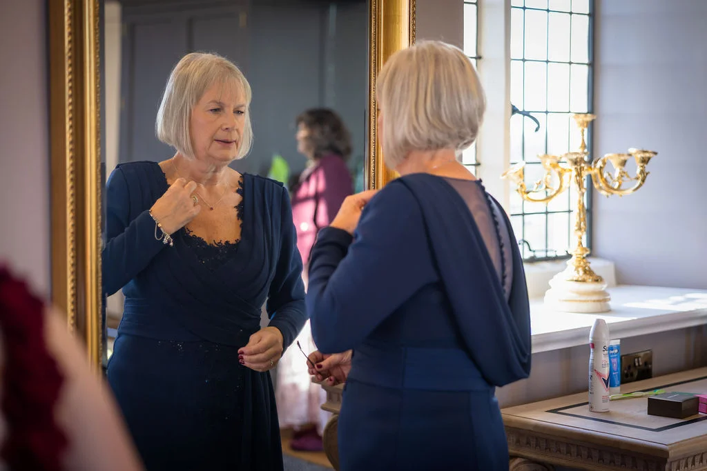 A mature bride with short grey hair adjusts her necklace while looking into a large ornate mirror during wedding preparations.