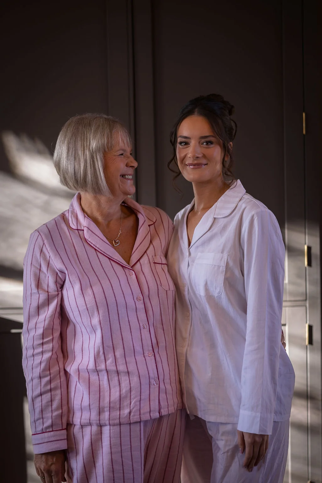 A smiling bride and her mother stand together wearing matching pyjamas during the morning bridal preparations bathed in soft natural light.