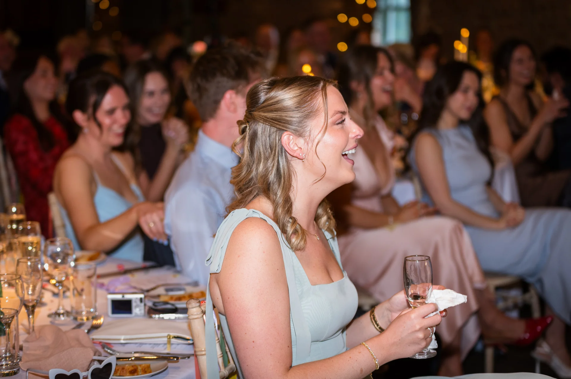 A wedding guest sits at a candlelit reception table, laughing joyfully while holding a glass of champagne during the speeches.