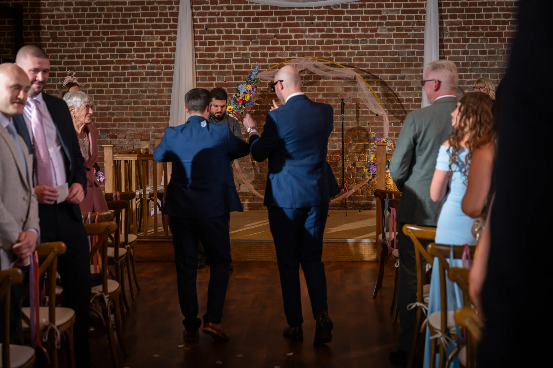 Groomsmen in blue suits walk down the aisle towards a circular floral arch inside a rustic brick barn wedding venue.