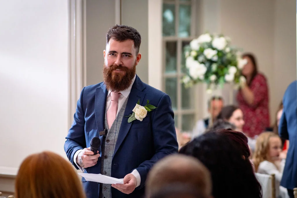 A bearded groom in a navy suit and tweed waistcoat delivers a speech holding a microphone during the wedding reception.