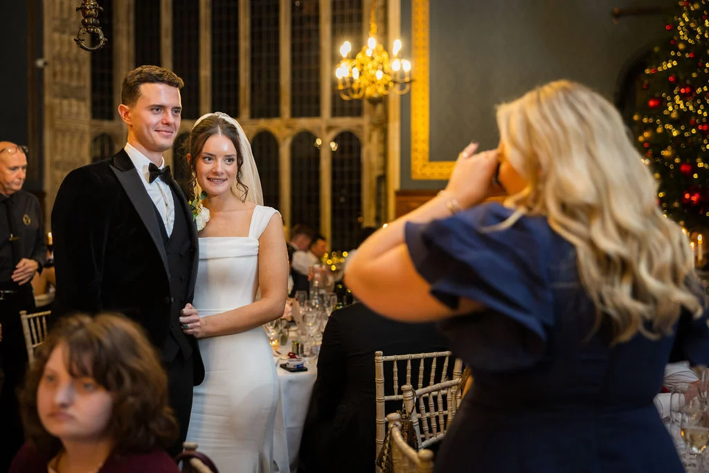 A bride and groom in formal attire pose smiling while a wedding guest takes a photo of them at the reception tables.