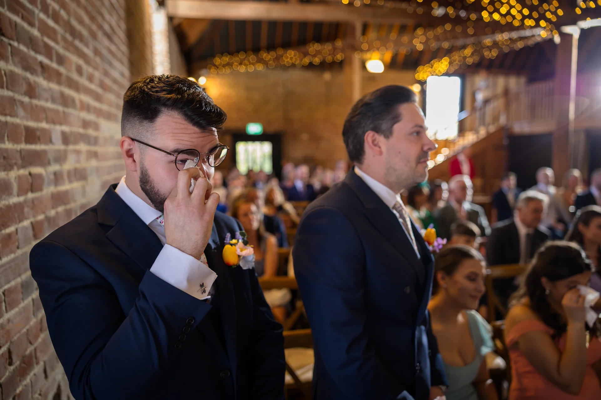 An emotional groom wearing glasses and a navy suit wipes away a tear during a rustic barn wedding ceremony while seated guests look on.