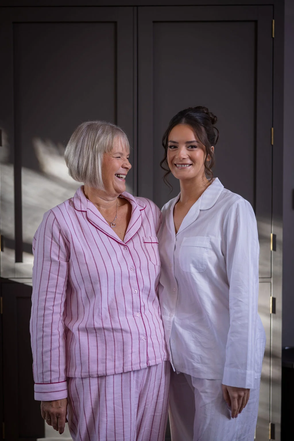 A smiling bride in white pyjamas stands happily next to the mother of the bride who is laughing while wearing matching pink striped pyjamas during morning preparations.