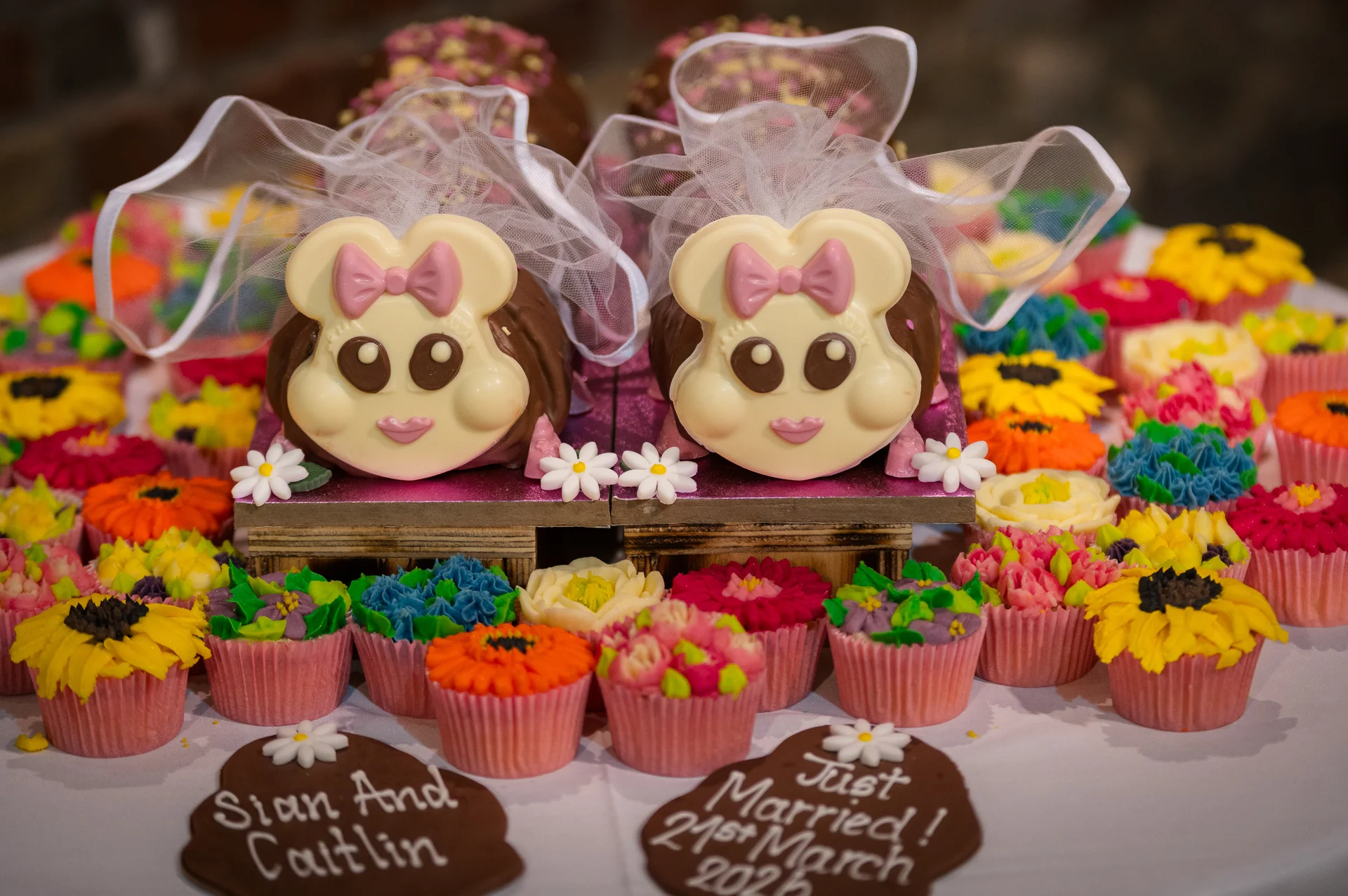 A whimsical dessert table features two novelty chocolate bride cakes adorned with white tulle veils, surrounded by a vibrant display of colourful floral cupcakes.