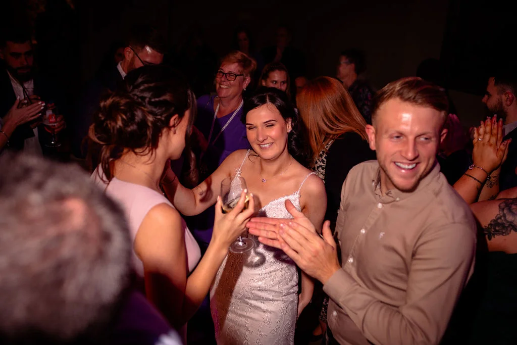 A joyful bride in a sparkly lace wedding dress dances with guests amidst a lively, crowded evening reception under moody lighting.