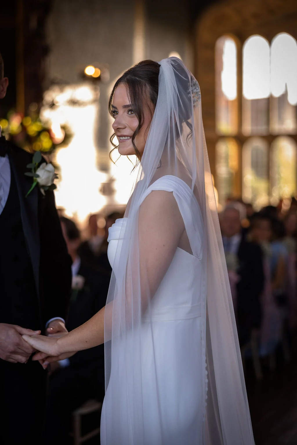 A smiling bride holding hands with her groom inside a sunlit church during the wedding ceremony, wearing a sleek crepe gown and a flowing veil.