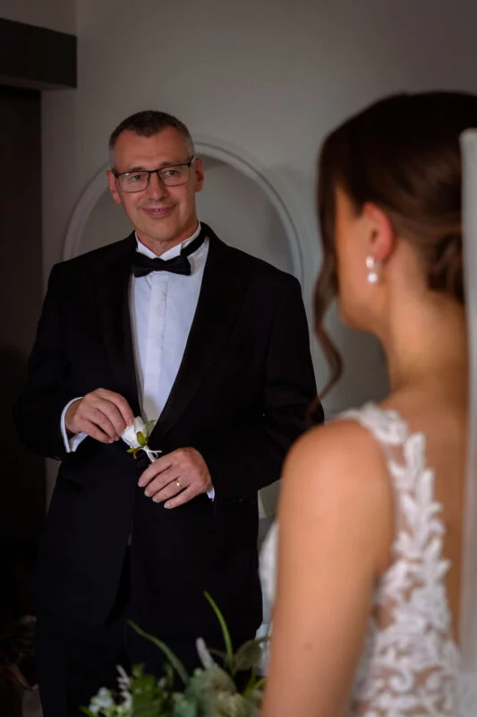 A proud father in a classic black tuxedo smiles tenderly at his daughter in her lace wedding dress during a first look moment.