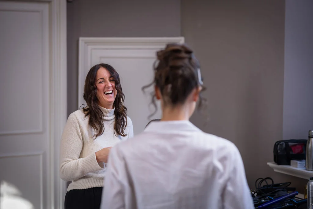 A smiling woman shares a joyful, laughing moment with a bride during wedding morning preparations indoors.