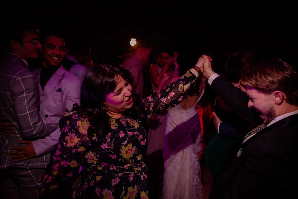 Wedding guests laughing and dancing energetically with raised arms on a lively evening reception dance floor.