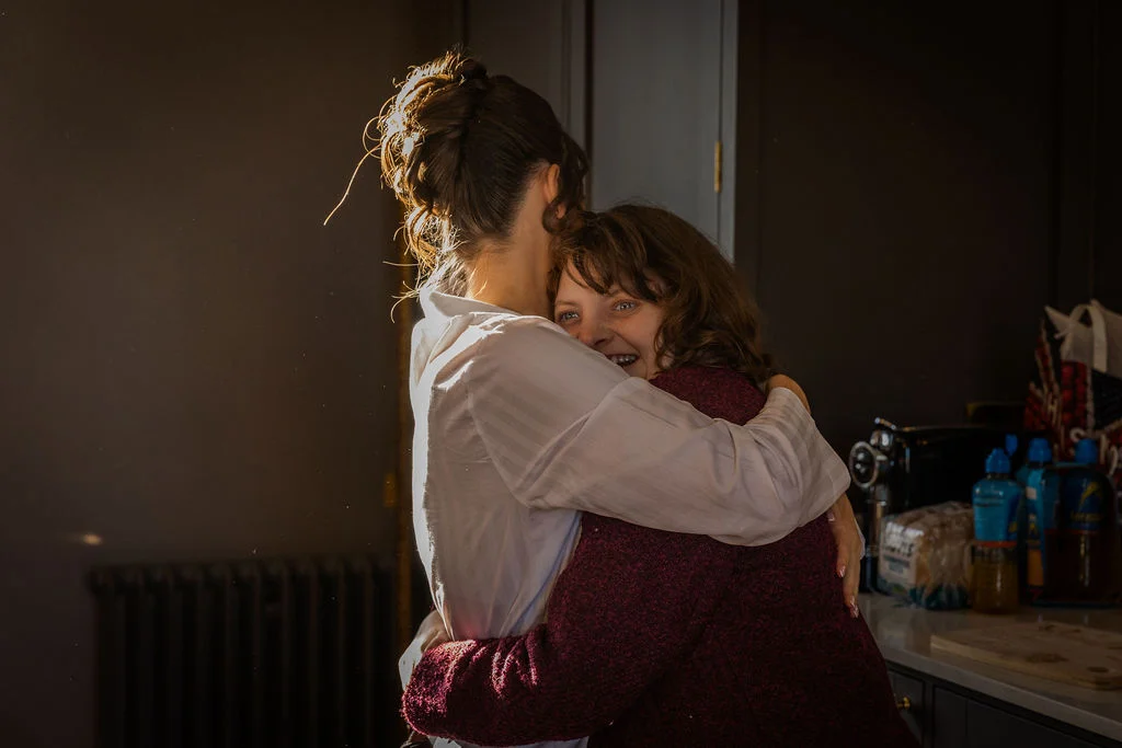 A tender moment captured in warm backlighting as two women embrace emotionally during morning bridal preparations.