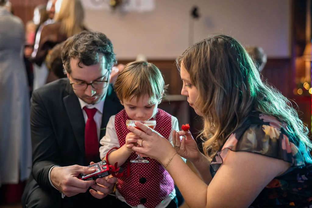 A young page boy in a red polka dot waistcoat sips a drink from a glass held by a woman, while a man looks on with a small red camera.