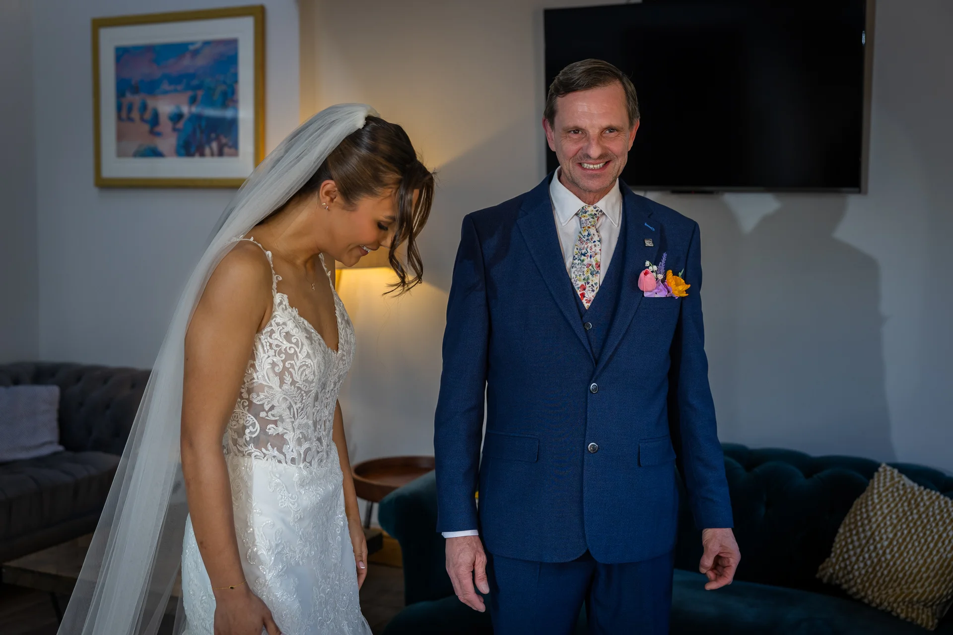 A smiling father in a navy suit and floral tie shares a tender first look moment with his daughter, who is wearing a lace wedding dress and veil.