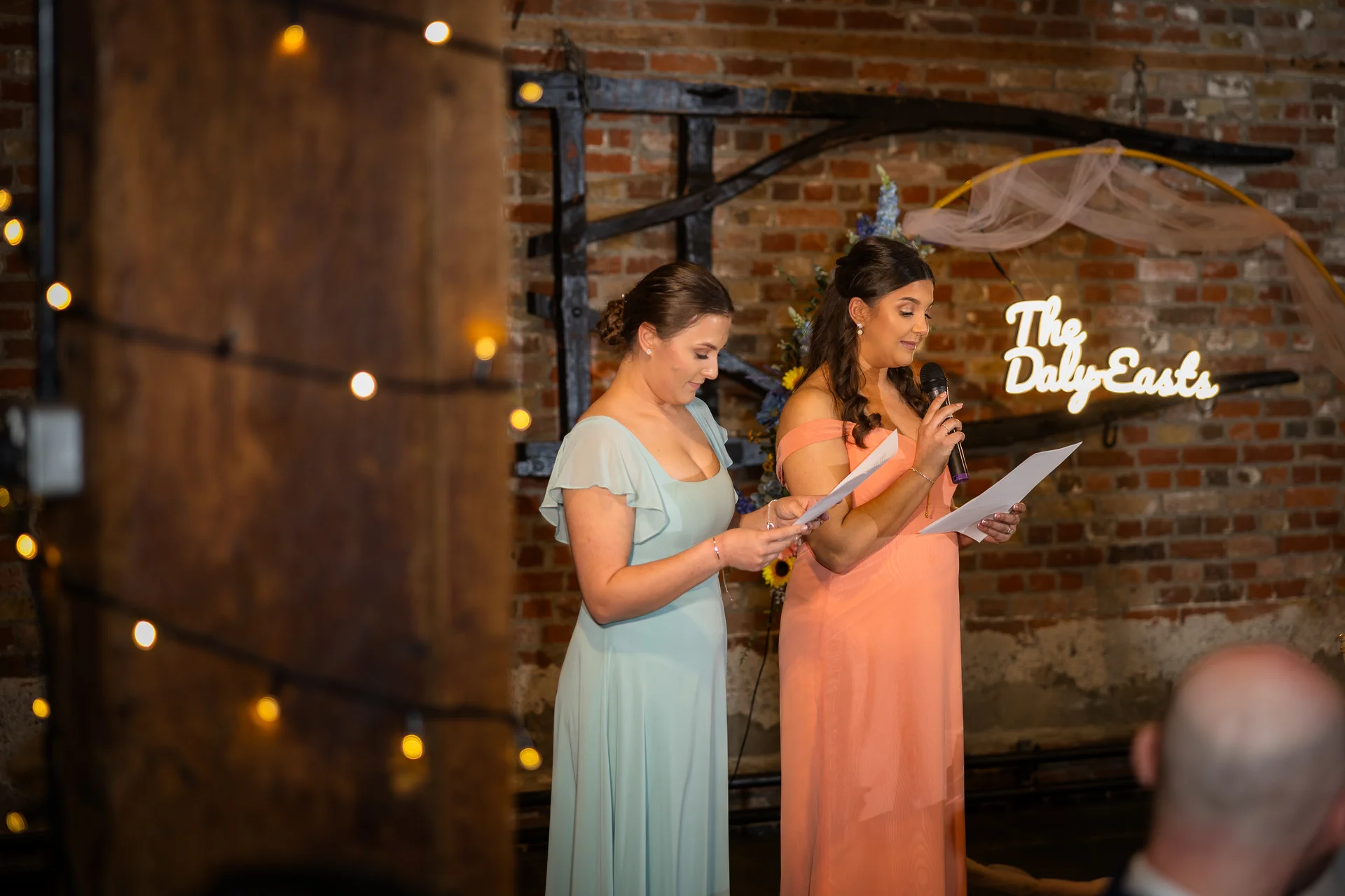 Two bridesmaids deliver speeches holding microphones and notes in front of an exposed brick wall adorned with a custom neon sign and fairy lights.