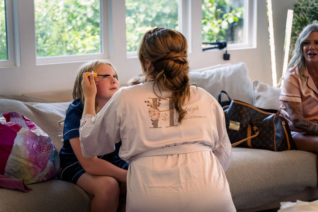 A person wearing a personalised bridal party robe applies makeup to a young girl seated on a sofa during morning wedding preparations.