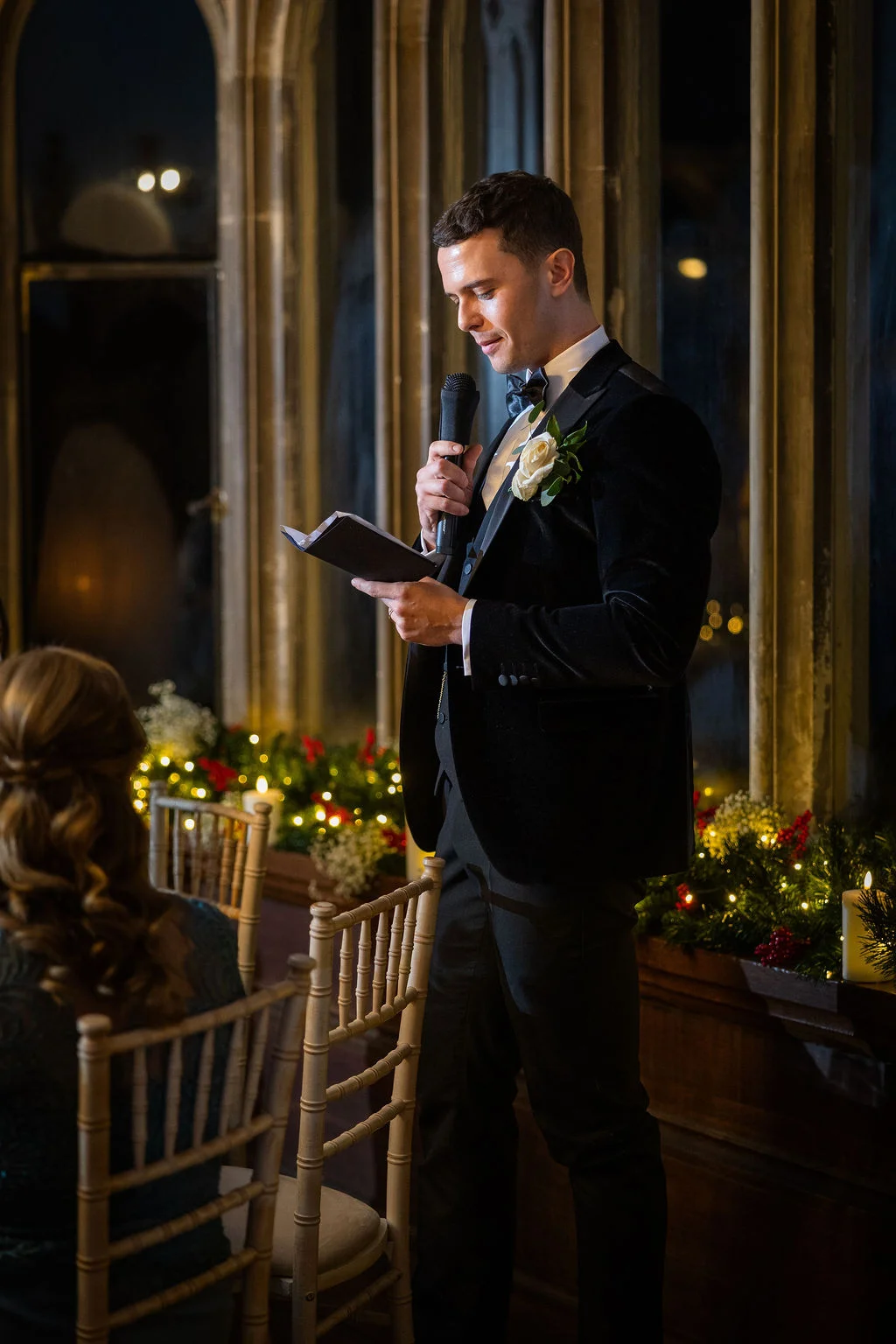 A groom in a black velvet tuxedo delivers his wedding speech indoors at night, holding a microphone and notes against a backdrop of tall windows and festive candlelight.