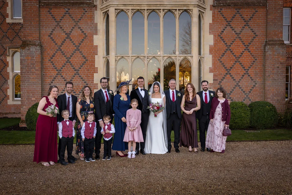 A large wedding party and family group pose together outside a historic red brick manor house with large stone-mullioned windows.