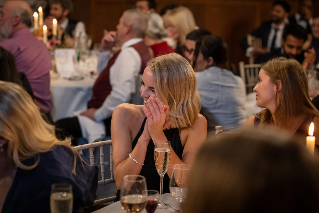 A blonde guest laughs joyfully with her hands clasped during the wedding breakfast speeches in a candlelit reception room.